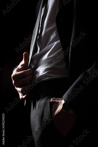 Elegant young fashion man in tuxedo hold tie in hands the other hands in pocket .on black background photo