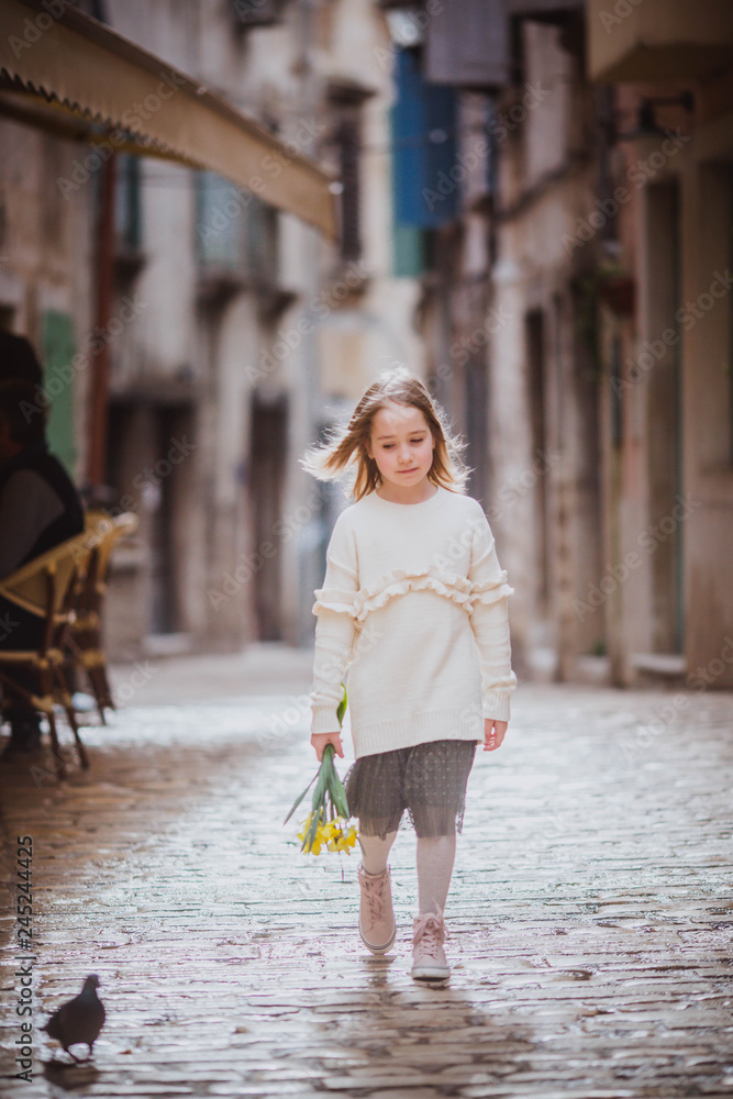 Adorable little girl in trend clothes standing in old town in sunny spring day with yellow flowers