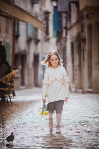 Adorable little girl in trend clothes standing in old town in sunny spring day with yellow flowers