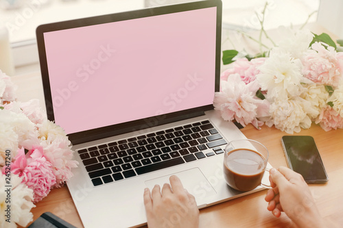 Girl hands on stylish laptop with empty screen and coffee, phone, black notebook and peonies on rustic wooden table. Freelance concept. Workplace. Space for text.  Women's Day.