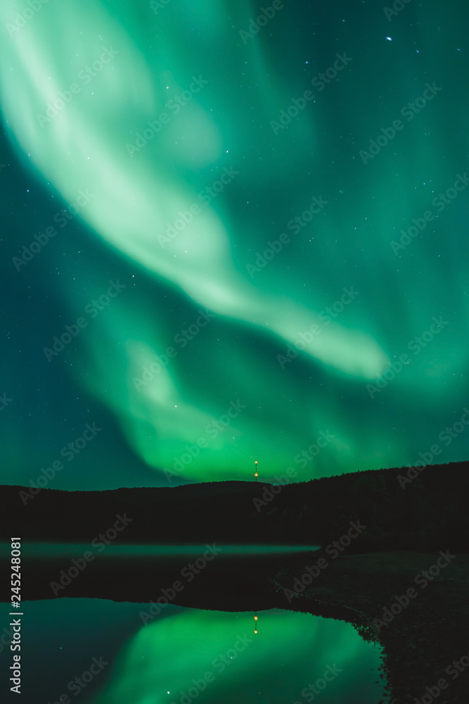 Fototapeta premium Green Northern Lights and stars in the night sky over the lake. Observing Aurora Borealis at North. Light phenomenon in the Arctic Circle. Abstract background with copy space. 