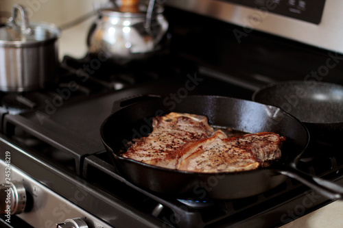 New York strip steaks frying in a cast iron pan on a natural gas stove top.