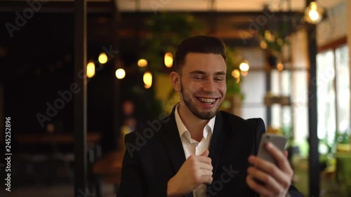 Young man is sitting in a cafe indoors, talking with somebody in a video chat, showing him his braces and pointing on them. Man is smiling and laughing out loud. photo