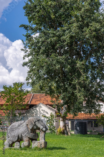 Belavadi, Karnataka, India - November 2, 2013: Veera Narayana Temple. Closeup of gray stone small statue of elephant set on green lawn in garden. Red roofs of neighboring houses under cloudscape. photo