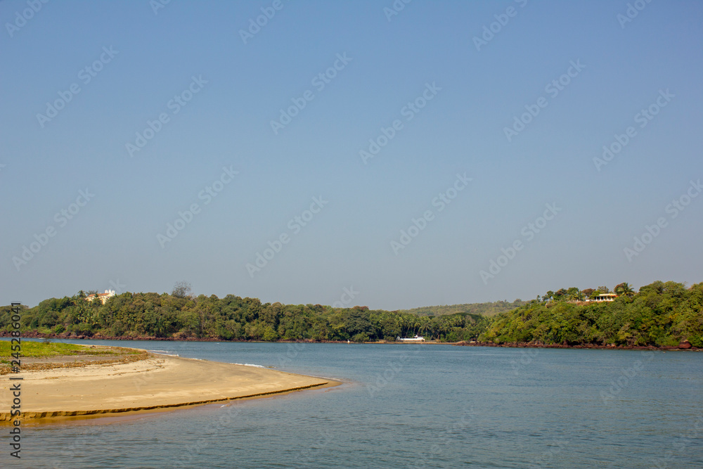 sandy shore on the background of the river and hills with green trees and houses under a clear blue sky