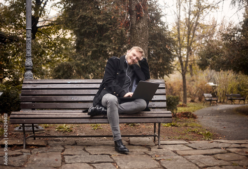 one young relaxed man, sitting on bench in public park, using laptop, looking to camera. Formal wear or smart casual. Full length shot.