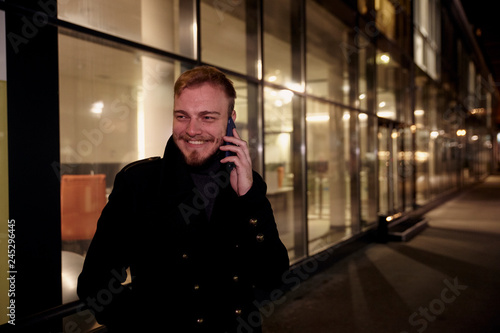 night time, one young smiling and happy man, 20-29 years, talking over his phone, standing in Autumn coat on street, in front of business offices windows outdoors.