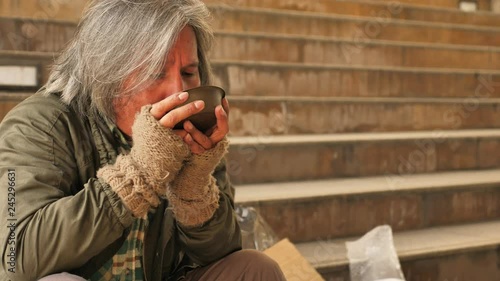 Homeless old man eating soup received from volunteers while sitting on stairs in the city