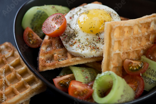 Close-up of waffles with fried egg and fresh vegetables served in a black bowl, selective focus