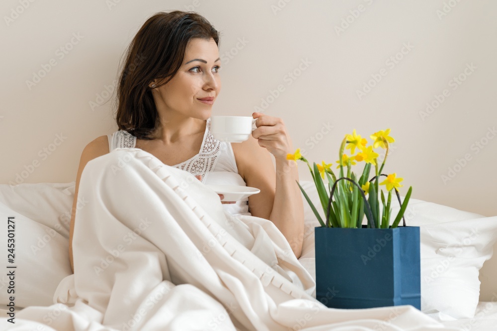 Happy smiling adult woman with bouquet of yellow flowers and cup of coffee sitting at home in bed Happy smiling adult woman with bouquet of yellow flowers and cup of coffee sitting at home in bed