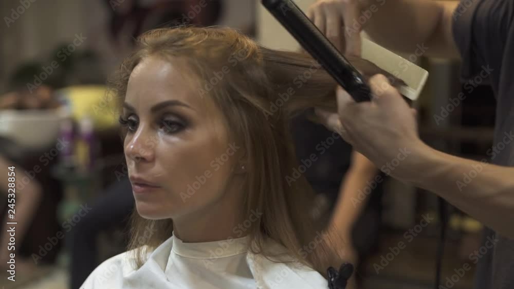 Hairstylist combing and straightening hair with hair iron in hairdressing salon. Hairdresser doing female hairstyle with hot tongs in barbershop. Beauty and style concept. Hairstylist combing and straightening hair with hair iron in hairdressing salon. Hairdresser doing female hairstyle with hot tongs in barbershop. Beauty and style concept.