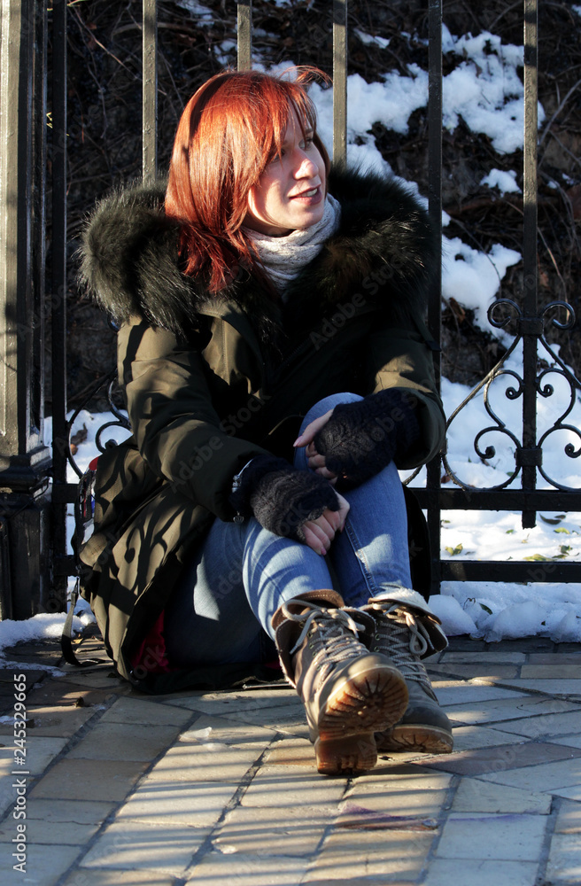A young girl sits on the floor, arms around her knees, smiles and looks away on a sunny winter day.