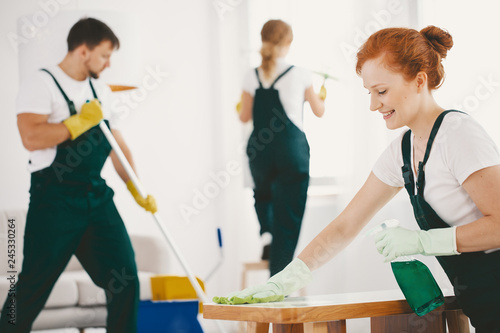 Smiling woman with gloves and spray wiping table while cleaning interior with crew