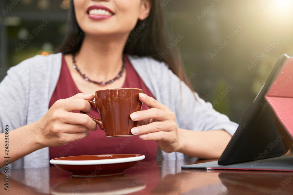 Close-up of smiling young lady sitting at the table with touchpad in front of her and holding cup of coffee Close-up of smiling young lady sitting at the table with touchpad in front of her and holding cup of coffee