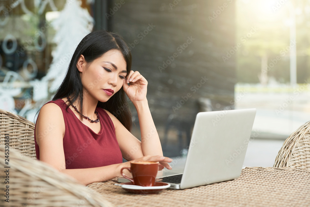 Serious Asian woman sitting at outdoor cafe drinking coffee and working online at laptop, she concentrating on her work Serious Asian woman sitting at outdoor cafe drinking coffee and working online at laptop, she concentrating on her work