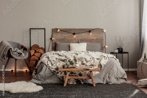 Trendy grey chair with blanket and log of wood next to cozy double bed with wooden headboard and light bulbs photo