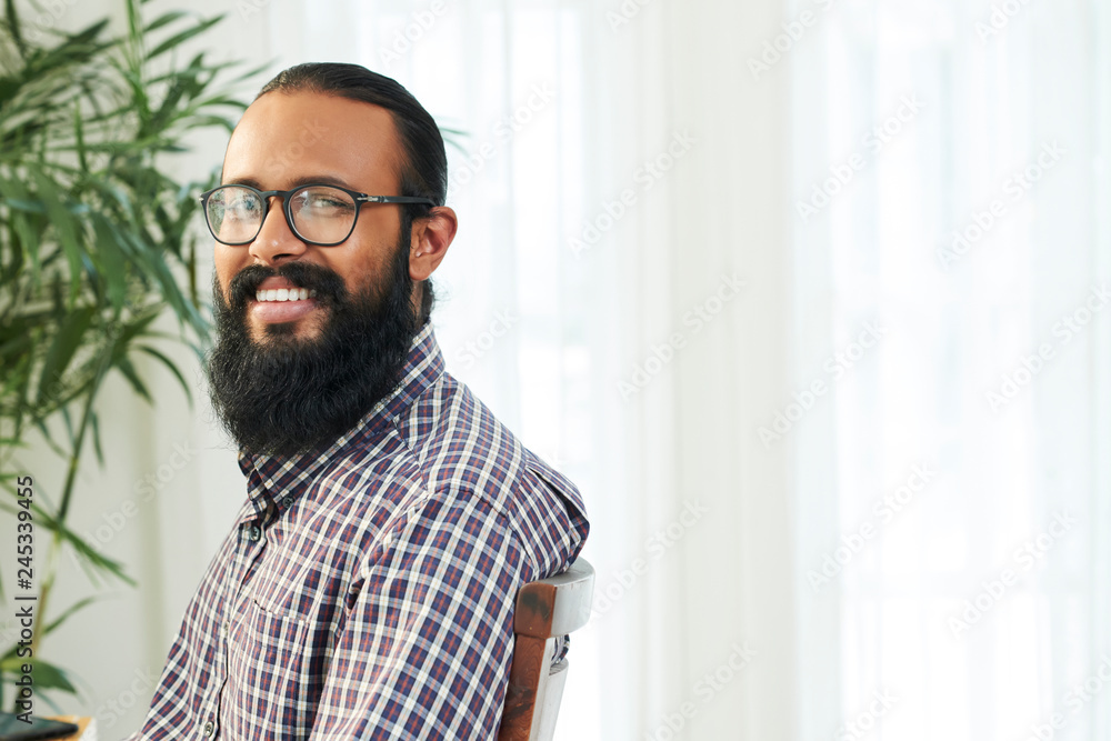 Happy young bearded businessman with toothy smile looking at you while sitting in office in front of camera Happy young bearded businessman with toothy smile looking at you while sitting in office in front of camera