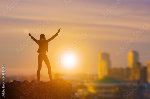 Slender girl businessman on top of a mountain holding her hand up  against the background of the city in the rays of the sun. Business concept idea  happiness  success and achievement  leadership.