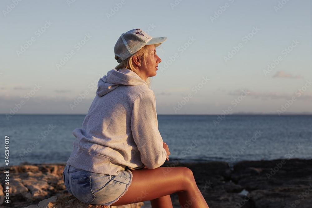 Thoughtful pensive young Caucasian woman has sporty body, dressed in casual hoodie and shorts, rests after jogging at seaside, has dark hair combed in plait, admires beautiful scenery on coastline.