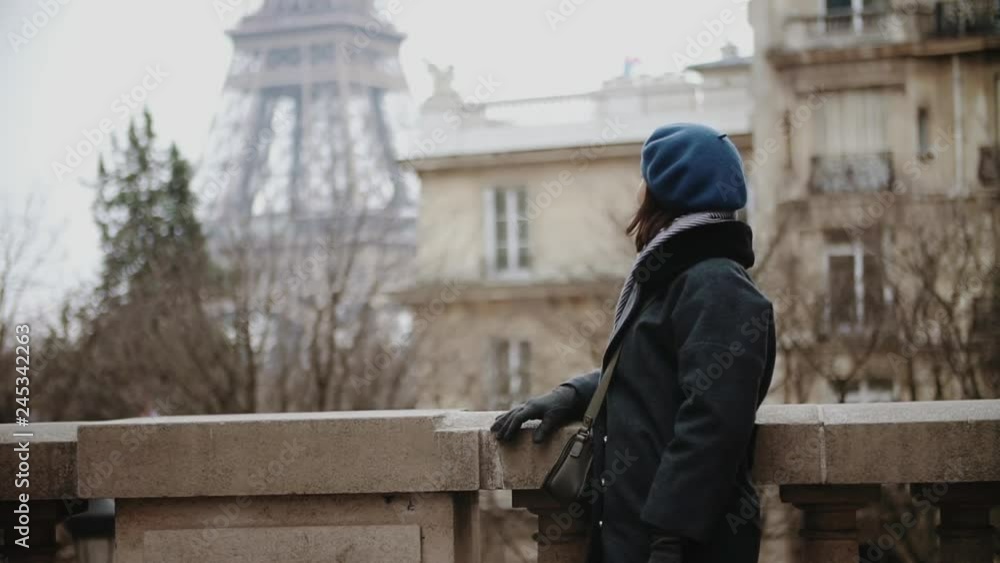 European stylish woman in beret and winter coat posing into the camera looking around and smiling chilling on the Camoens Avenue street by the Eiffel tower in Paris, France European stylish woman in beret and winter coat posing into the camera looking around and smiling chilling on the Camoens Avenue street by the Eiffel tower in Paris, France