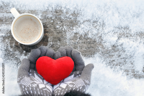 Valentine's day and Love frosty picture: a Cup of hot coffee on a wooden bench and hands in knitted gray gloves hold a plush red heart on a winter day. Valentine day