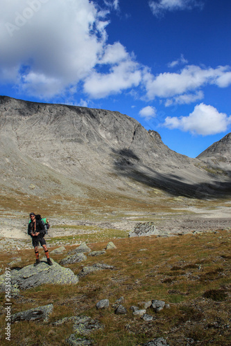 Young white Caucasian male tourist with backpack against the backdrop of mountains and clear blue sky with clouds in Karelia, Russia