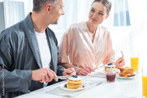 selective focus of beautiful couple in robes during breakfast with pancakes and orange juice in kitchen