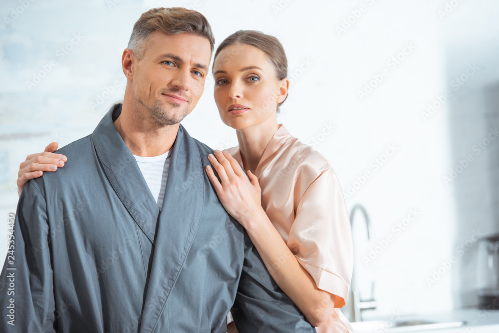 beautiful woman in robe looking at camera and embracing man in kitchen beautiful woman in robe looking at camera and embracing man in kitchen