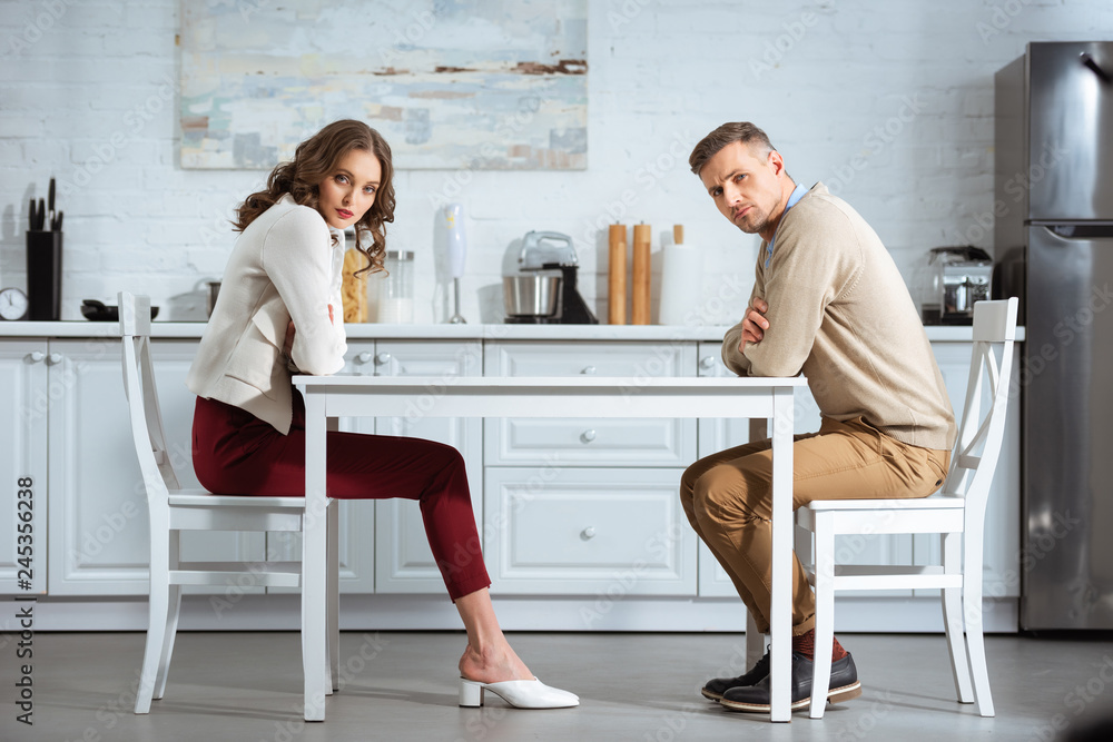 dissatisfied couple sitting with arms crossed at table in kitchen dissatisfied couple sitting with arms crossed at table in kitchen