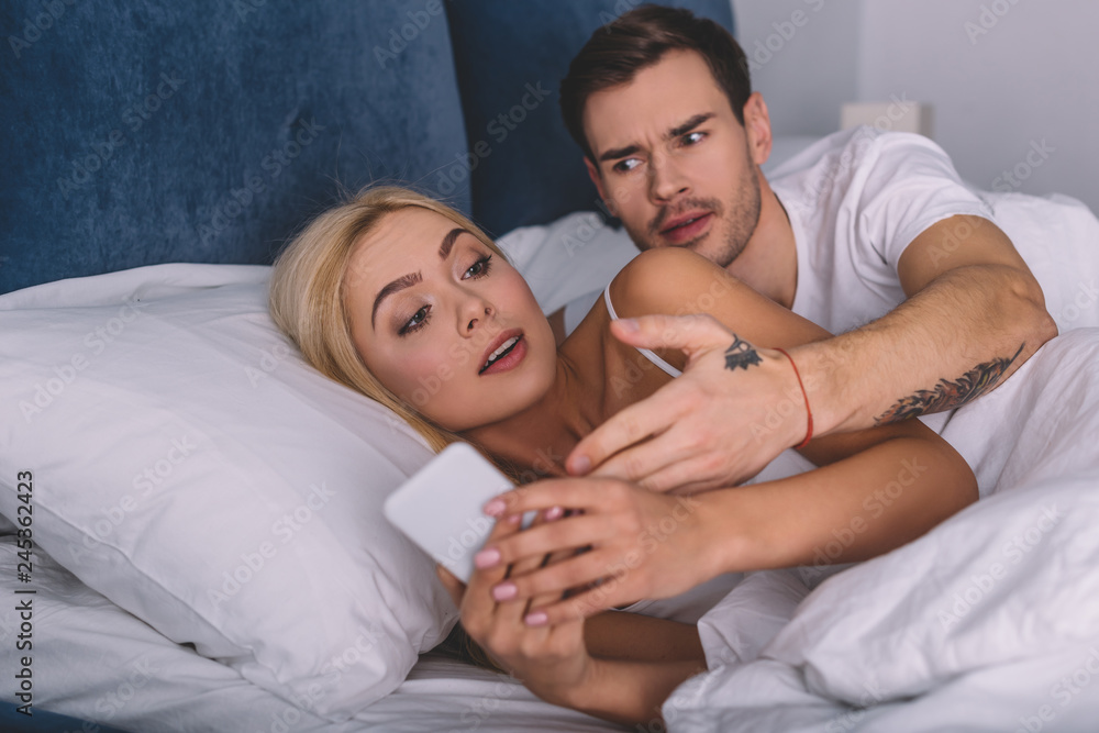 angry man looking at suspicious girlfriend using smartphone in bed, mistrust concept angry man looking at suspicious girlfriend using smartphone in bed, mistrust concept