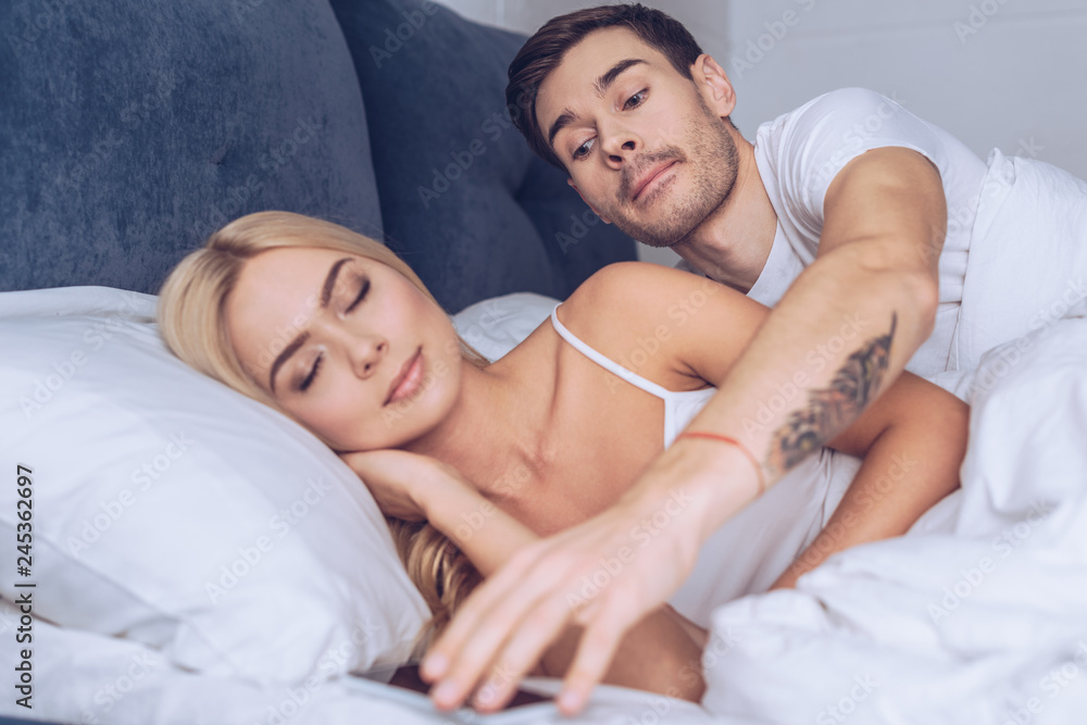 suspicious young man reaching for smartphone while beautiful girlfriend sleeping in bed, mistrust concept suspicious young man reaching for smartphone while beautiful girlfriend sleeping in bed, mistrust concept