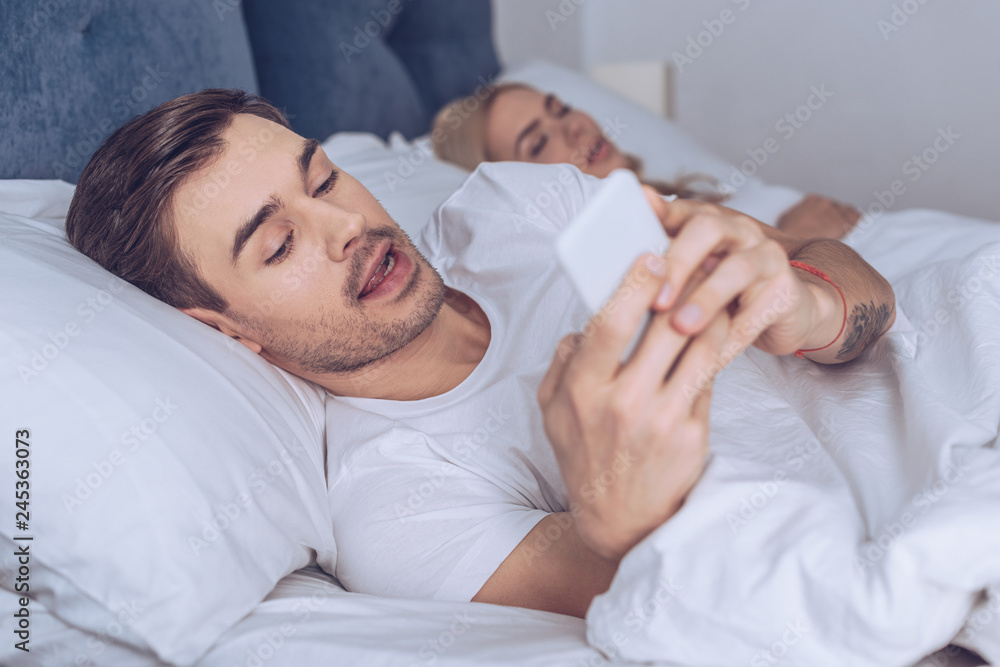 suspicious young man using smartphone while lying with sleeping wife in bed suspicious young man using smartphone while lying with sleeping wife in bed