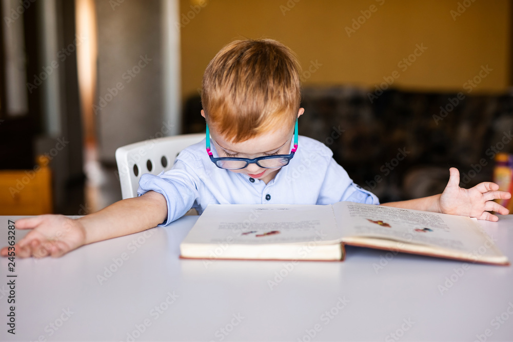 Naklejka premium Cute toddler boy with down syndrome with big glasses reading intesting book