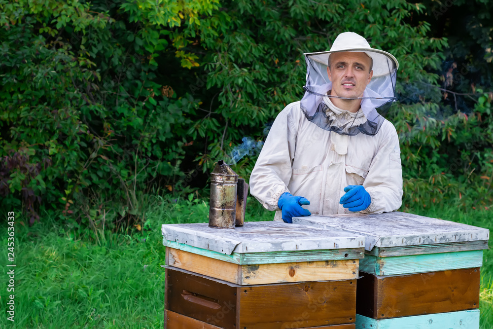 Portrait of a young beekeeper in an apiary near a hive in a protective suit. Beekeeping concept. Beekeeper harvesting honey. Portrait of a young beekeeper in an apiary near a hive in a protective suit. Beekeeping concept. Beekeeper harvesting honey.