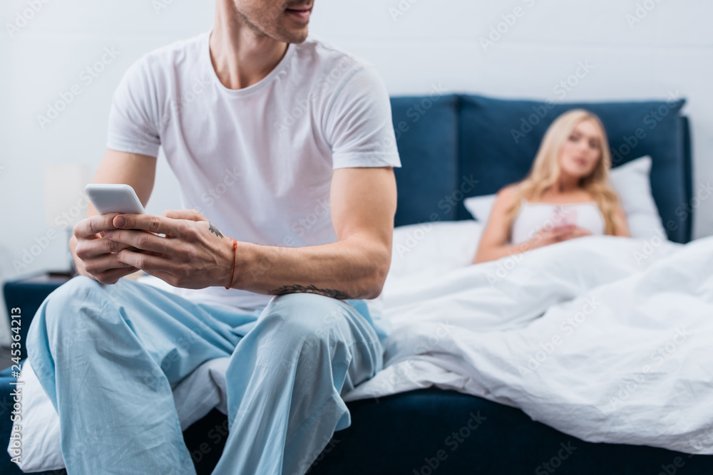 Fototapeta premium cropped shot of young man in pajamas using smartphone while jealous wife lying in bed behind