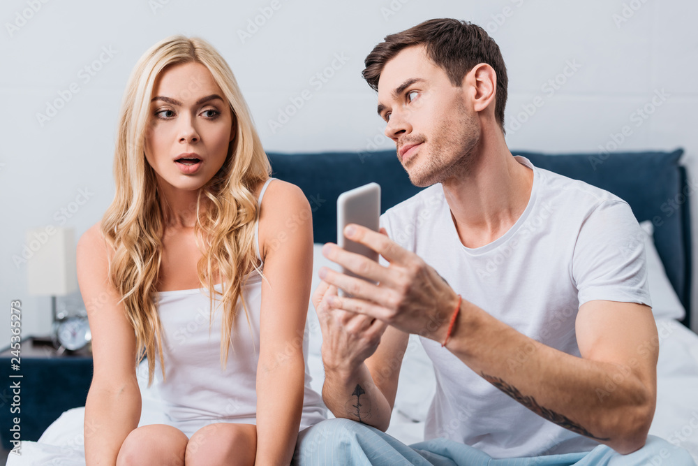 jealous man showing smartphone to confused girlfriend sitting on bed, distrust concept jealous man showing smartphone to confused girlfriend sitting on bed, distrust concept
