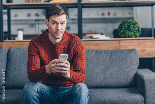 suspicious young man holding smartphone and looking at camera while sitting on couch at home photo
