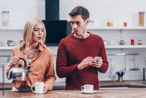 Fotografi jealous young woman holding kettle and looking at husband with smartphone in kit