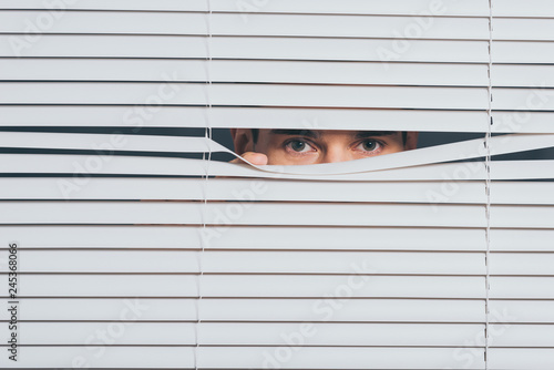 suspicious young man peeking and looking at camera through blinds, mistrust concept photo