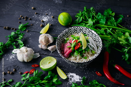 Pho Bo vietnamese soup with noodles, beef, chili pepper, onion, cilantro and lime on dark black background