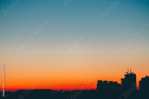 Cityscape with wonderful varicolored vivid dawn. Amazing blue sky with orange sunny light above dark silhouettes of city buildings. Atmospheric background of warm sunrise. Copy space.