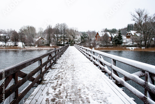 Wooden bridge in winter, trees and and wooden houses covered by snow in a village © Michele Ursi