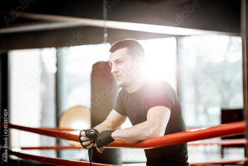 Portrait of a young athletic man in black sportswear standing on the boxxing ring at the gym