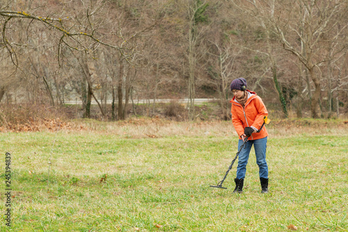 a girl in an orange jacket with a metal detector in his hands, is on the field in search of antiquity