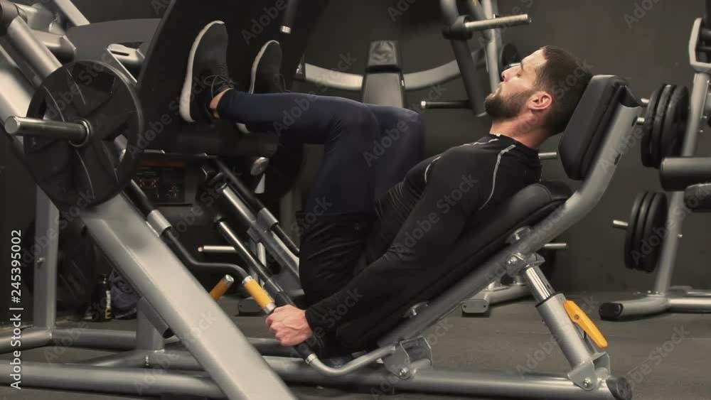 Side view of athletic handsome young man with beard in black sportswear exercising on leg press machine in gym. Long shot, static shot. Training concept. Side view of athletic handsome young man with beard in black sportswear exercising on leg press machine in gym. Long shot, static shot. Training concept.