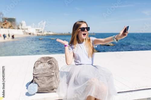 Young pretty girl with long hair is sitting on pier. She wears lush skirt with gray T-shirt. She is taking selfie photo. © Look!