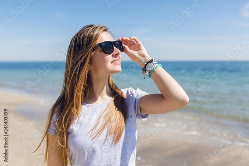 Close-up portrait of a young pretty girl with long hair  walking on the beach near sea. She wears gray T-shirt and some ornamentation on hand. She is smiling to the sun. © Look!