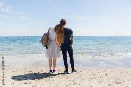 Young couple is standing near sea. He wears black T-shirt and pants. She has long hair, gray T-shirt, skirt and bag. She put head on his shoulder. View from back.