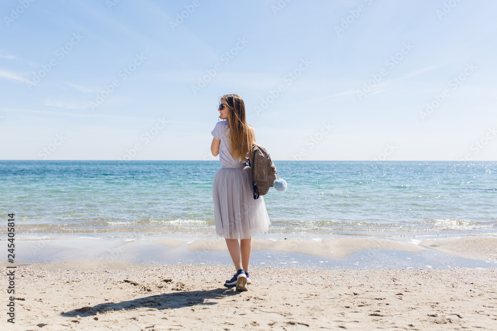 Young girl with long hair and bag on back is standing near sea. She wears lush gray skirt and gray T-shirt with sneakers. View from back. Young girl with long hair and bag on back is standing near sea. She wears lush gray skirt and gray T-shirt with sneakers. View from back.