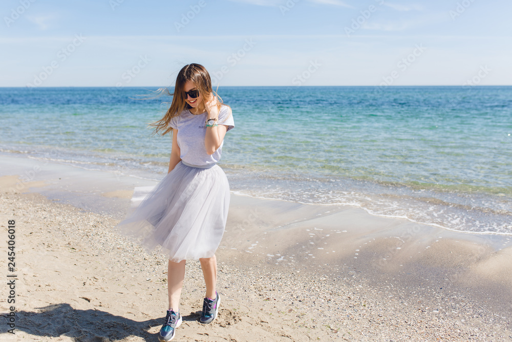 Young girl with long hair is walking near blue sea. She wears lush gray skirt and gray T-sirt with sneakers. Young girl with long hair is walking near blue sea. She wears lush gray skirt and gray T-sirt with sneakers.